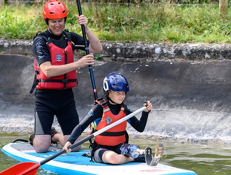 A male instructor and a boy with a bionic leg paddle through the water on top of a board